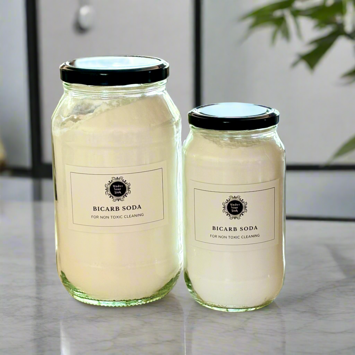 Two clear glass jars, one larger (2Litres) and one smaller (1litre). They both have simple white label with Under your Sink Logo and description Bicarb- all round Eco-cleaner – mild strength alkali. They are filled with white crystalline Bicarb granules inside. Background is of a kitchen and its a clean image to show green cleaning. This is a product image for website as this is for sale in various sizes, online with Australia wide delivery.