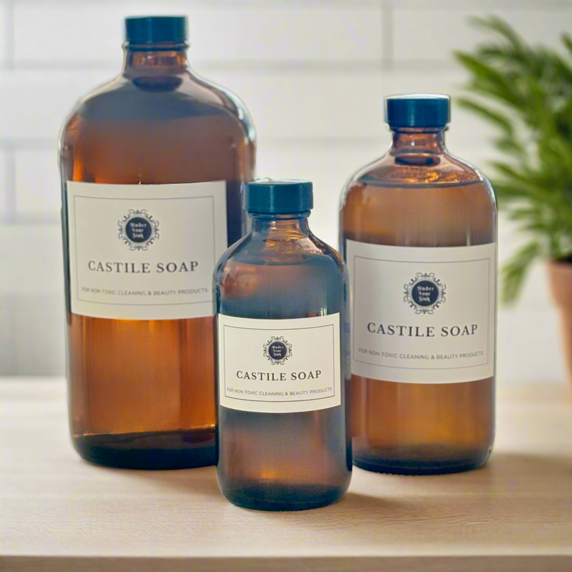 A collection of 3 amber glass bottles in 1litre, 500ml and 250ml sizes. Each has a simple white label with Under your Sink Logo and description Castile Soap – For non toxic cleaning and beauty products. The bottles are set in a kitchen with a blurred background for effect. It is a product image for website as this is for sale in various sizes, online with Australia wide delivery.