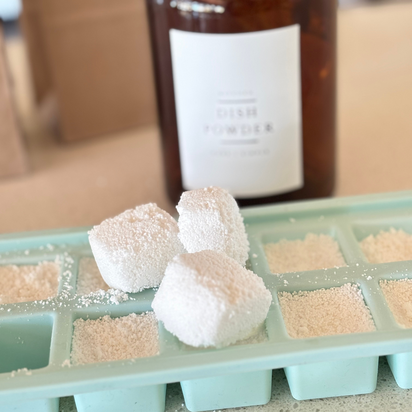 An ice cube tray with handmade dishwasher tablets using Under your Sink Dish Powder. In background is a vintage amber glass apothecary jar